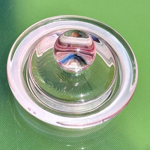 May include: A clear glass lid with a pinkish hue, featuring a rounded knob at the center. The lid has a circular design with a raised edge, and the knob has a striped pattern. The object is likely a decorative or functional item.