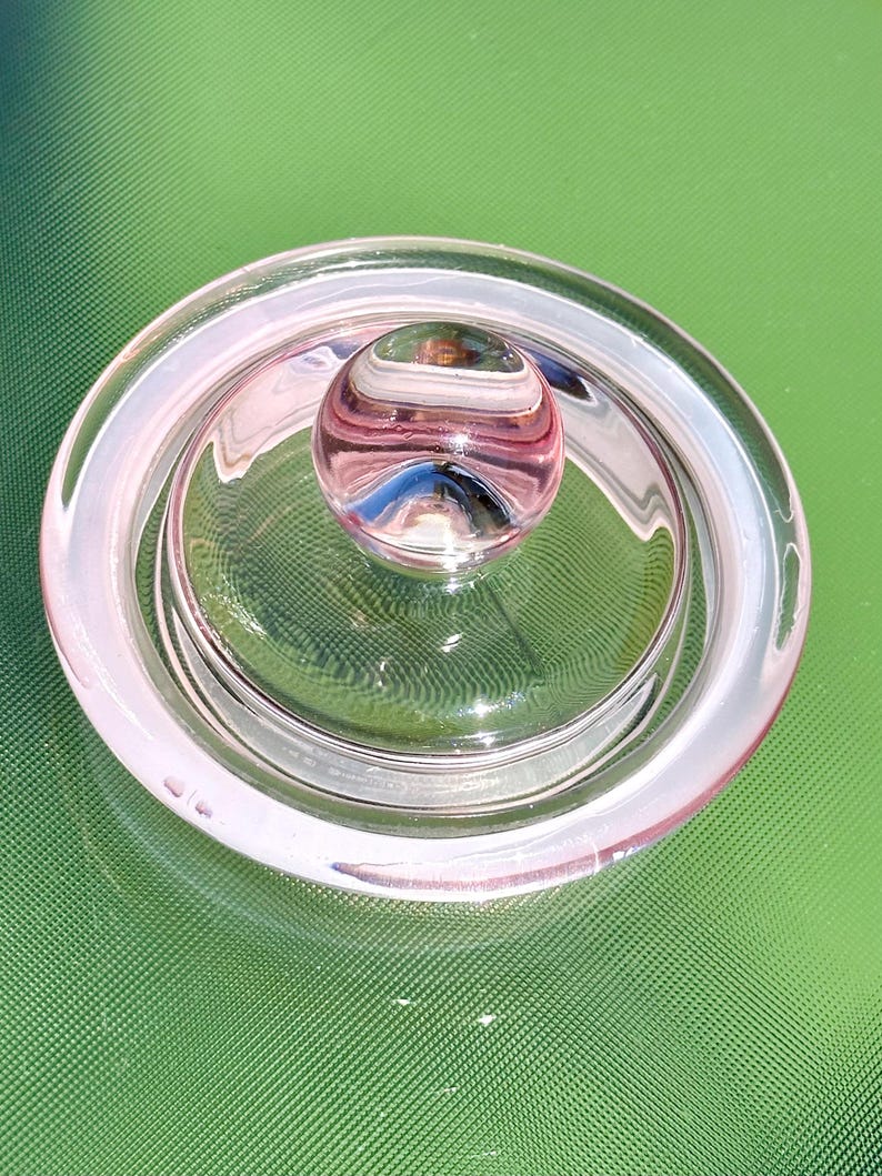 May include: A clear glass lid with a pinkish hue, featuring a rounded knob at the center. The lid has a circular design with a raised edge, and the knob has a striped pattern. The object is likely a decorative or functional item.