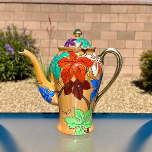 May include: A ceramic teapot with a glossy peach-colored base, featuring a vibrant hand-painted pattern of multi-colored grape leaves in shades of red, green, blue, and white. It includes a matching lid with a gold-toned finial and a gold handle.