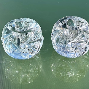 May include: Two clear, cut-crystal round bowls with intricate starburst and leaf patterns. The bowls are reflecting on a green surface, creating a mirrored effect. The crystal bowls are likely decorative items.