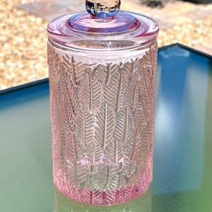 May include: A pink glass jar with a lid and a round knob. The jar has a textured feather design. The jar is sitting on a glass table. The background is blurred.