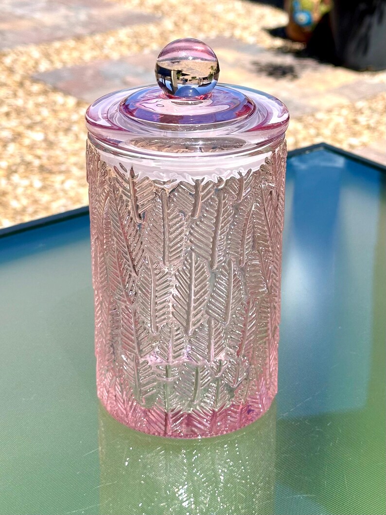 May include: A pink glass jar with a lid and a round knob. The jar has a textured feather design. The jar is sitting on a glass table. The background is blurred.