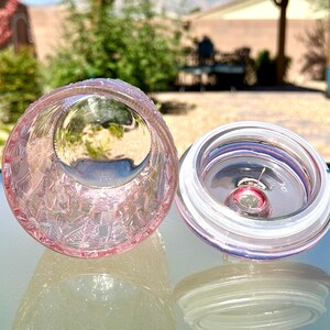 May include: A pink glass jar with a textured exterior and a clear lid. The jar is open, revealing the interior. The lid is off to the side, showing a clear seal. The jar is sitting on a reflective surface.