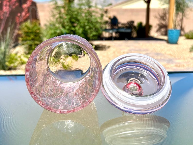 May include: A pink glass jar with a textured exterior and a clear lid. The jar is open, revealing the interior. The lid is off to the side, showing a clear seal. The jar is sitting on a reflective surface.