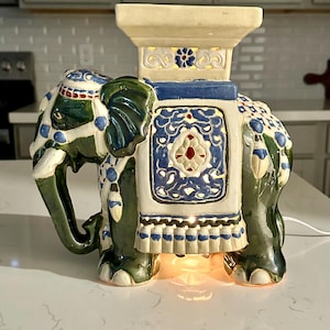 May include: A ceramic elephant figurine with a cream-colored platform on its back. The elephant is painted in shades of green, blue, and white with intricate floral and geometric patterns. The platform has a blue and white floral design. The elephant is lit from within.