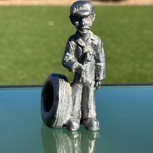 May include: A silver-colored metal figurine of a mechanic wearing a cap and overalls, holding a wrench. The mechanic is standing next to a tire. The figurine is on a reflective surface.