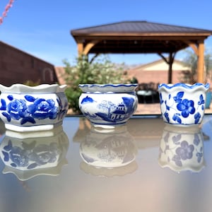 May include: Three small, white ceramic pots with blue floral designs. The pots have scalloped edges and are arranged on a reflective surface. The designs include roses, a landscape scene, and stylized flowers. The background features a gazebo and greenery.