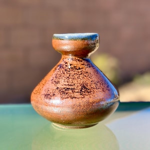 May include: A small, handcrafted ceramic vase with a bulbous body and a narrow neck. The vase features a speckled brown glaze with hints of blue and green around the rim. The vase is approximately 4 inches tall.