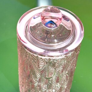 May include: A pink glass jar with a lid and a round knob. The jar has a cylindrical shape with a textured, leaf-like pattern. The lid is circular and the knob is a small, round sphere. The jar is sitting on a green surface.