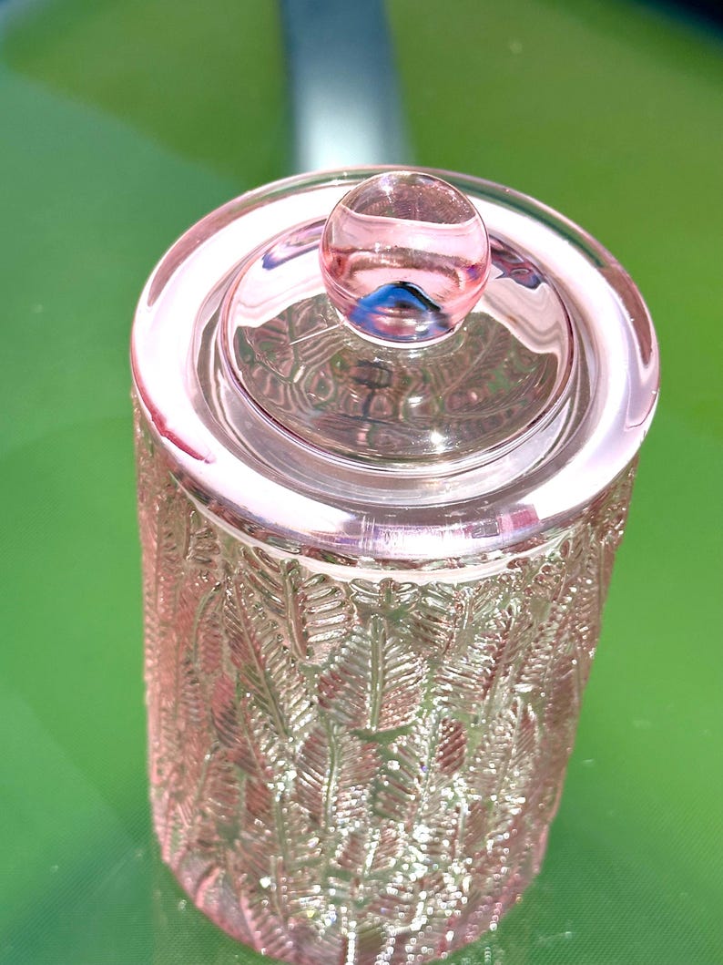 May include: A pink glass jar with a lid and a round knob. The jar has a cylindrical shape with a textured, leaf-like pattern. The lid is circular and the knob is a small, round sphere. The jar is sitting on a green surface.