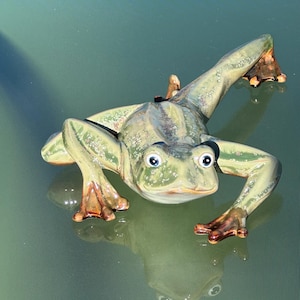 May include: A ceramic frog figurine in shades of green and brown, with large blue eyes. The frog is posed as if leaping forward, with its legs extended. The figurine has a glossy finish and is reflected in the surface below.