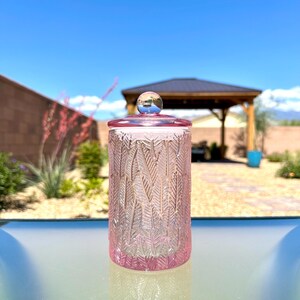 May include: A pink glass jar with a lid. The jar has a textured feather design and a clear glass lid with a small blue ball on top. The jar is sitting on a glass surface, with a blue sky and a gazebo in the background.