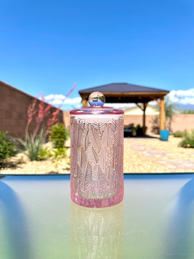 May include: A pink glass jar with a lid. The jar has a textured feather design and a clear glass lid with a small blue ball on top. The jar is sitting on a glass surface, with a blue sky and a gazebo in the background.