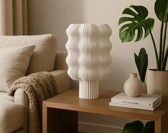 Könnte beinhalten: Eine weiße, strukturierte Keramikvase mit wellenförmigem Design, die auf einem Holztisch steht. Die Vase hat eine zylindrische Form mit einem Sockel. Eine kleine, runde Vase und Bücher befinden sich ebenfalls auf dem Tisch, mit einem Sofa und Pflanzen im Hintergrund.