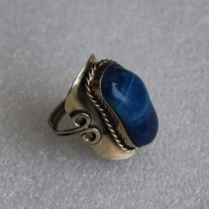 Könnte beinhalten: Silberring mit einem großen, ovalen blauen Stein. Der Stein ist in einer silbernen Lünette mit einem gedrehten Seildesign eingefasst. Der Ring hat dekorative Wirbel an den Seiten.