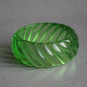 Könnte beinhalten: Ein leuchtend grünes Glasarmband mit Spiralmuster. Das Armband ist ein massives Band aus transparentem grünem Glas mit diagonalen Rillen, die ein strukturiertes Muster erzeugen. Ein auffälliges und stilvolles Accessoire.