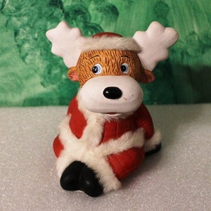May include: A ceramic reindeer figurine with a red and white Santa Claus-style outfit. The reindeer has a white face, black nose, and blue eyes. It is wearing a red hat with white trim and has white antlers. The figurine is sitting down.
