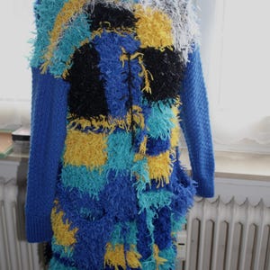 Könnte beinhalten: Ein lebendiger, strukturierter Mantel in Blau-, Gelb-, Schwarz- und Weißtönen. Das langärmlige Kleidungsstück hat ein Patchwork-Design mit einer zotteligen, fransigen Textur. Der Mantel wird auf einer Schaufensterpuppe präsentiert.