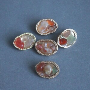 Könnte beinhalten: Fünf ovale Edelstein-Cabochons mit goldfarbenen, strukturierten Fassungen. Die Edelsteine weisen eine Mischung aus Farben auf, darunter Rot, Orange, Weiß und Grün. Die Cabochons sind auf einer grauen Oberfläche angeordnet.