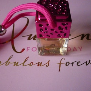 May include: A pink and gold car air freshener with a decorative metal cage and a pink string hanging from it. The air freshener is sitting on a pink surface with the text "fabulous forever" printed on it.