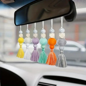 May include: Seven colourful car charms with wooden beads and tassels. Each charm features a different colour felt ball and a white wooden bead. The charms are hanging from a rear view mirror.