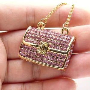 May include: A gold-tone chain necklace with a pink rhinestone handbag pendant. The handbag is detailed with a gold-tone clasp.