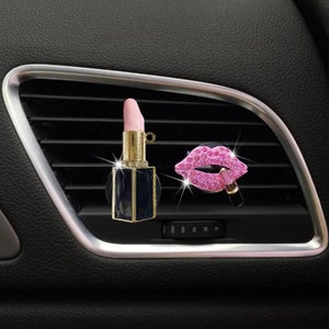 May include: Two car air fresheners. One is a lipstick-shaped ornament with a pink lipstick top and a black base. The other is a pink jeweled lips design with a lipstick. Both are placed on a car vent.