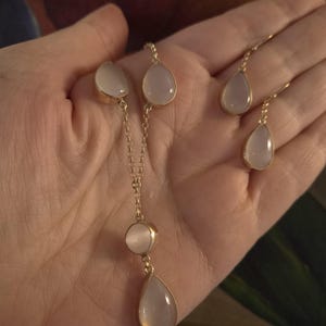 May include: A gold-toned jewelry set consisting of a necklace and a pair of drop earrings. The necklace features a delicate chain with two teardrop-shaped stones and one circular stone. The earrings feature matching teardrop-shaped stones.