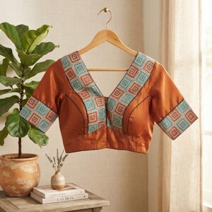May include: A rust-colored blouse featuring a deep V-neckline and short sleeves, accented with a geometric pattern in shades of teal, orange, and white along the neckline and sleeve cuffs. The garment is displayed on a wooden hanger.