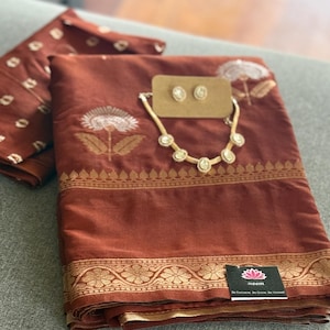 Könnte beinhalten: Ein gefalteter rostfarbener Stoff mit goldener Bordüre und Blumenmuster, weißen Blumenstickereien sowie einer passenden Halskette und Ohrringen auf einer Karte. Ein kleines schwarzes Etikett mit rosa Lotus-Symbol und dem Text Bloom.