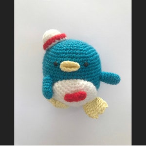 Pode incluir: Um brinquedo de pinguim de crochê azul e branco com um chapéu vermelho e branco. O pinguim tem um bico amarelo e olhos pretos.