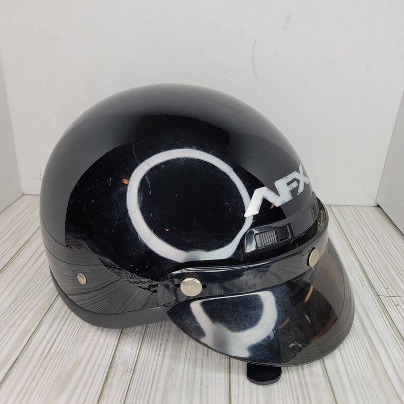 May include: A black motorcycle helmet with a visor. The helmet features the white text "AFX" on the side. The visor is made of a dark, transparent material. The helmet has a rounded shape and is designed for head protection.