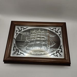 Puede incluir: Espejo enmarcado con un grabado detallado de un velero. El barco está representado con líneas intrincadas, navegando sobre el agua. El marco es de color marrón oscuro, con anclas decorativas en las esquinas.