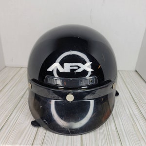 May include: Black motorcycle helmet with a visor. The helmet features the white letters "NFX" inside a white circle on the front. The visor is black and has two silver-coloured snaps. The helmet is glossy and has some scratches.