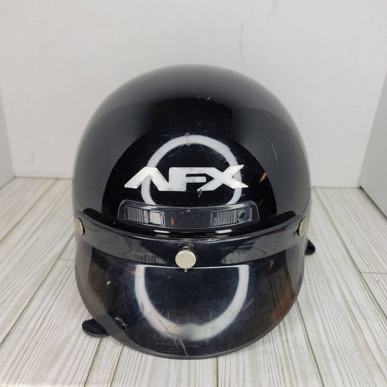 May include: Black motorcycle helmet with a visor. The helmet features the white letters "NFX" inside a white circle on the front. The visor is black and has two silver-coloured snaps. The helmet is glossy and has some scratches.