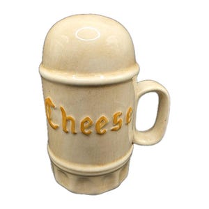May include: A beige ceramic cheese shaker with a handle. The word "Cheese" is written in yellow, raised letters on the side. The shaker has a rounded top and a hexagonal base.