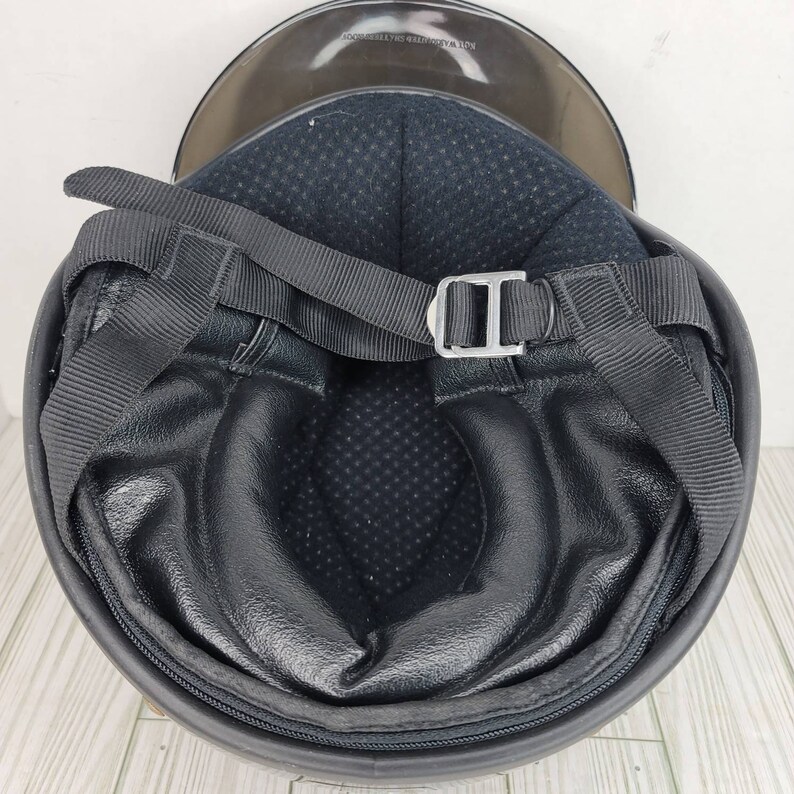 May include: Black motorcycle helmet with a clear visor. The interior has black padding and a black strap with a silver buckle. The helmet is designed for safety and protection.