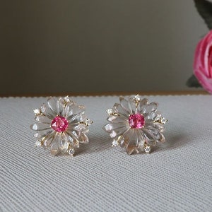 May include: A pair of delicate earrings featuring a clear crystal flower design with a pink gemstone center. The earrings are set in gold.