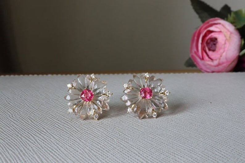 May include: A pair of delicate earrings featuring a clear crystal flower design with a pink gemstone center. The earrings are set in gold.