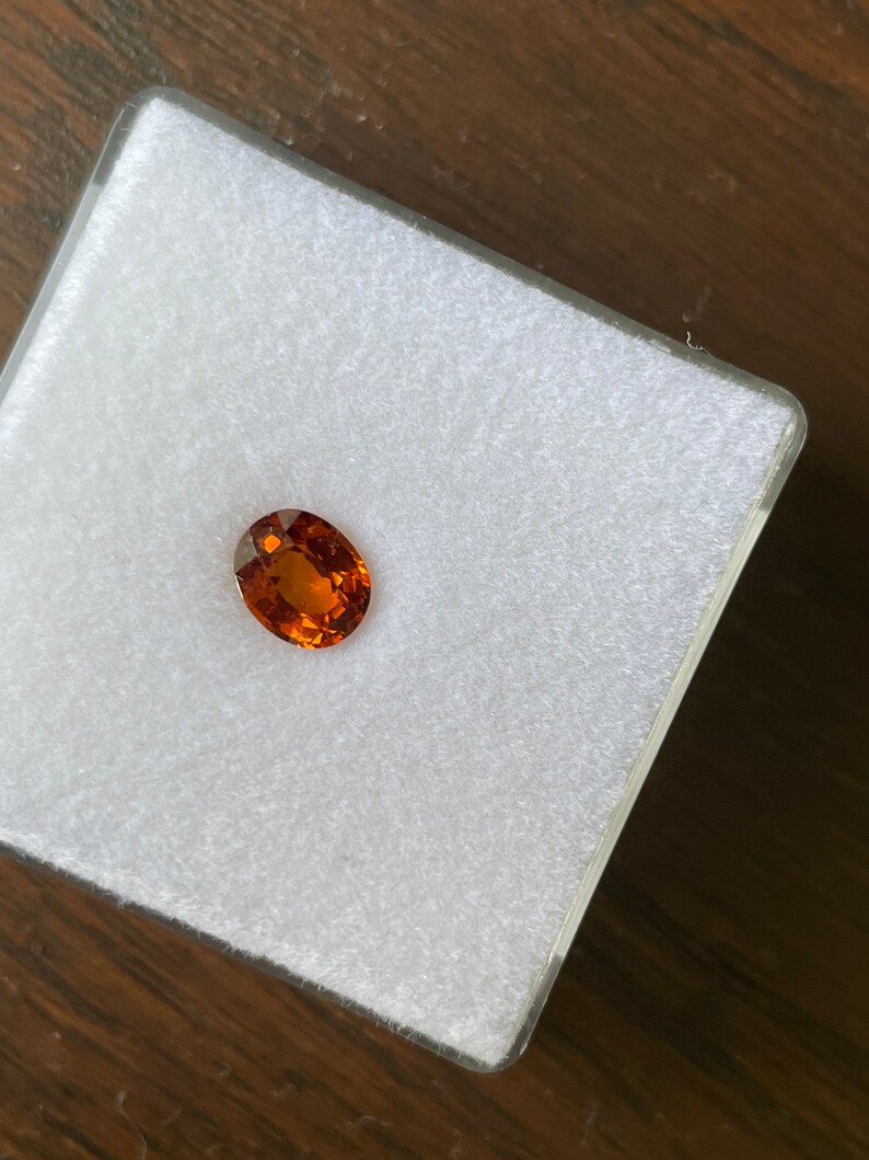 May include: A close-up of a small, oval-shaped, orange gemstone in a white, velvet-lined box.