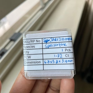 May include: A small, clear plastic box with a white label containing information about a Spessartine gemstone. The label includes the SKU/RP number, species, quantity, total carat weight (TCW), and dimensions. The gemstone is a Spessartine, weighing 1.32 carats and measuring 6.8 x 5.8 x 3.4 millimeters.