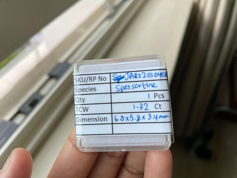 May include: A small, clear plastic box with a white label containing information about a Spessartine gemstone. The label includes the SKU/RP number, species, quantity, total carat weight (TCW), and dimensions. The gemstone is a Spessartine, weighing 1.32 carats and measuring 6.8 x 5.8 x 3.4 millimeters.