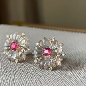 May include: A pair of delicate earrings featuring a pink gemstone set in a clear, carved flower design with small diamonds around the edges.
