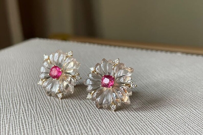 May include: A pair of delicate earrings featuring a pink gemstone set in a clear, carved flower design with small diamonds around the edges.