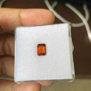 May include: A small, rectangular, orange-brown gemstone in a white, square, plastic box.