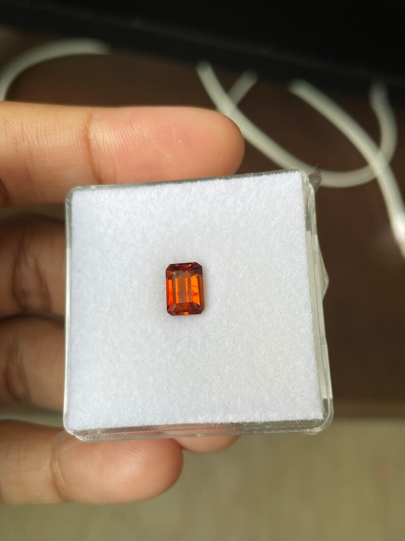 May include: A small, rectangular, orange-brown gemstone in a white, square, plastic box.