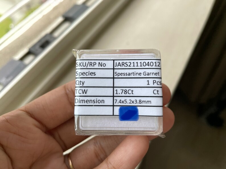 May include: A white label with black text listing the details of a 1.78 carat Spessartine Garnet gemstone. The label includes the SKU/RP number, species, quantity, total carat weight (TCW), and dimensions. The gemstone is 7.4 mm x 5.2 mm x 3.8 mm.