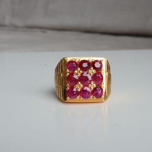 Könnte beinhalten: Ein goldfarbener Ring mit quadratischer Fassung, besetzt mit neun runden, tiefroten Edelsteinen. Der Ring hat ein strukturiertes Band und eine polierte Oberfläche, was einen klassischen und luxuriösen Look erzeugt. Die Edelsteine sind in einem Raster angeordnet.