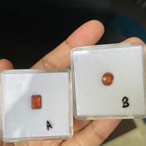 May include: Two small, clear plastic boxes with orange gemstones inside. The boxes are labeled "A" and "B".