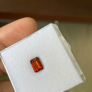 May include: A rectangular, orange gemstone in a white, square, plastic box.